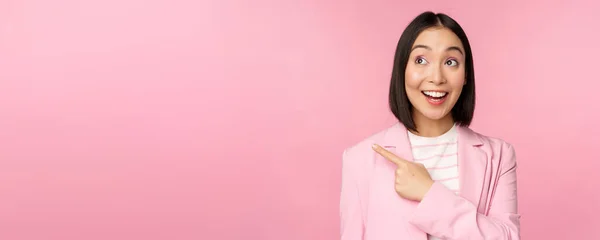 Enthusiastische junge asiatische Geschäftsfrau, Büroangestellte zeigt mit dem Finger nach links, blickt auf Banner, Werbung mit glücklichem Lächeln, zeigt Werbung, rosa Hintergrund — Stockfoto