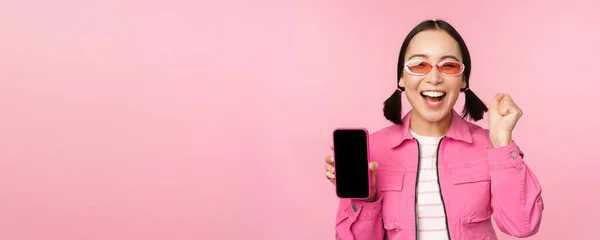 Возбужденная азиатка смеется и улыбается, показывает экран мобильного телефона, приложение для смартфона, стоя на розовом фоне — стоковое фото