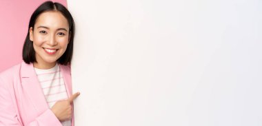 Mutlu gülümseyen Japon kadın portresi tahtayı işaret ediyor, boş alanda smth gösteriyor, pembe arka planda takım elbise içinde duruyor.