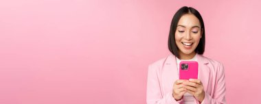 Akıllı telefonlu Asyalı bir kızın portresi. Gülümsüyor ve mutlu görünüyor. Pembe stüdyo arka planında duruyor.