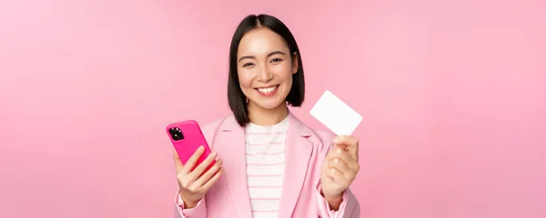 Зображення усміхненої щасливої азіатської бізнес-леді, що показує кредитну картку, що платить онлайн за додатком смартфона, замовлення з мобільним телефоном, стоїть на рожевому фоні — стокове фото