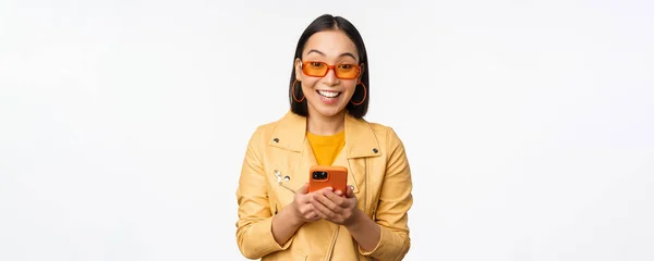 Nowoczesna azjatycka dziewczyna w okularach przeciwsłonecznych za pomocą telefonu komórkowego, uśmiechnięta i szczęśliwa, pozująca na białym tle — Zdjęcie stockowe