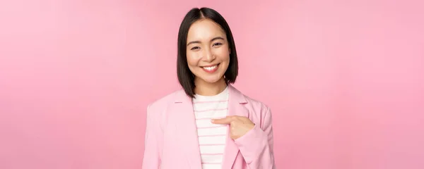 Junge Berufstätige. Lächelnde asiatische Geschäftsfrau, Verkäuferin zeigt mit dem Finger auf sich und sieht selbstbewusst aus, posiert im Anzug über rosa Hintergrund — Stockfoto