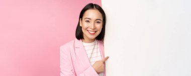 Mutlu gülümseyen Japon kadın portresi tahtayı işaret ediyor, boş alanda smth gösteriyor, pembe arka planda takım elbise içinde duruyor.