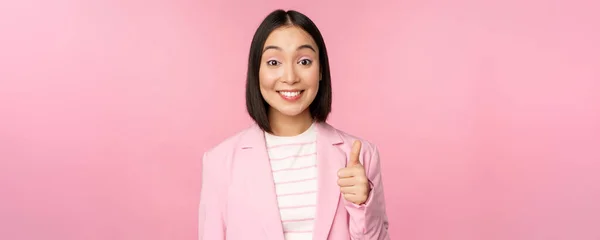 Retrato de mulher de negócios asiática sorrindo satisfeito, mostrando polegares para cima, louvor, como e aprovar, de pé em terno sobre fundo rosa — Fotografia de Stock