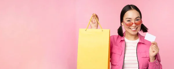 Boldog fiatal ázsiai nő mutatja hitelkártya és bevásárló táska, bolt eladó hirdetés, vásárlás smth a boltban, pózol a rózsaszín háttér — Stock Fotó