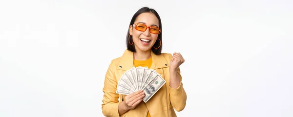 Elegante chica asiática sonriente sosteniendo dinero en efectivo, mostrando dólares y celebrando, de pie sobre fondo blanco — Foto de Stock