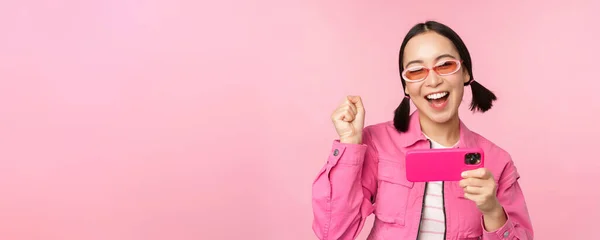 Счастливая улыбающаяся корейская девушка побеждает на мобильном телефоне, смотрит на горизонтальный экран смартфона и радуется, достигает цели, празднует, стоя на розовом фоне — стоковое фото