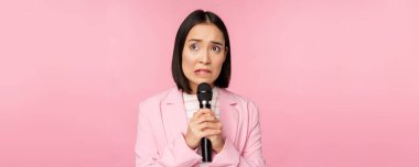 Tedirgin Asyalı kadın, herkesin içinde konuşuyor mikrofonla konferans veriyor, korkmuş görünüyor, pembe arka planda duruyor.