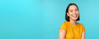 Covid-19 'dan aşı kampanyası. Mutlu ve sağlıklı Asyalı kız Coronavirüs aşısından sonra gülüyor, omzunda bandaj, sarı tişört, mavi arka plan