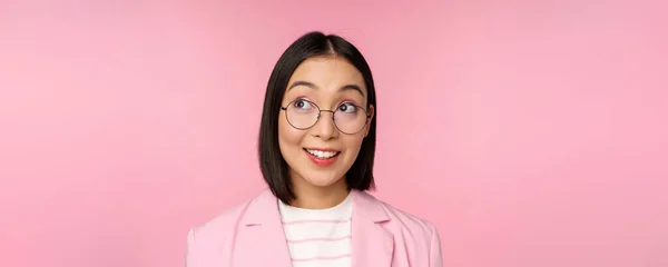 Enthusiastic saleswoman, asian businesswoman in glasses, looking intrigued at upper left corner logo, company advertisement, standing over pink background — Stock Photo, Image