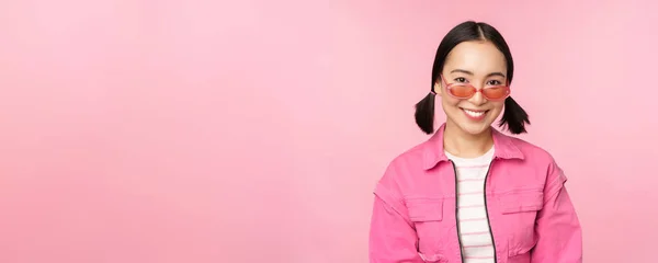 Close up de belo modelo feminino asiático em óculos de sol elegantes, posando contra fundo rosa na roupa da moda, espaço cópia — Fotografia de Stock