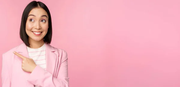 Entuzjastyczna młoda azjatycka bizneswoman, pracownik biurowy wskazujący palcem w lewo, patrząc na baner, reklama ze szczęśliwym uśmiechem, pokazując reklamę, różowe tło — Zdjęcie stockowe