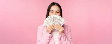 Takım elbiseli mutlu Asyalı kadın elinde para, mutlu yüz ifadesi olan dolarlar, pembe arka planda duruyor.