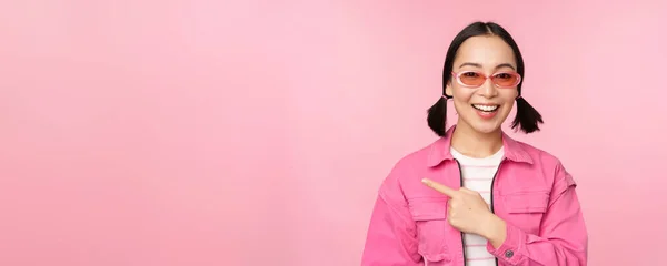 Retrato de sorrindo ásia menina no elegante roupa, óculos de sol, apontando dedo esquerdo, mostrando anúncio, banner, pé sobre rosa fundo — Fotografia de Stock