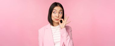 Asyalı kurumsal bir kadının portresi, ağzı mühürlü, dudakları kapalı anahtar hareketi, söz ver sır tut, pembe arka planda duran takım elbiseli.