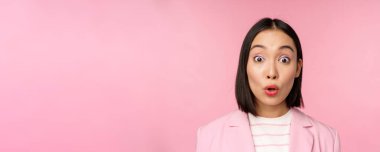 Asyalı şirket kadınının yakın portresi, iş kadını kameraya şaşırmış ve şaşırmış görünüyor, pembe arka plana karşı takım elbise içinde duruyor.