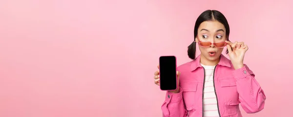 Snygg koreansk tjej, ung kvinna i solglasögon visar smartphone skärm, mobiltelefon app gränssnitt eller hemsida, står över rosa bakgrund — Stockfoto