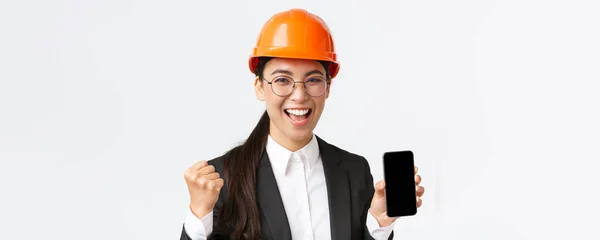 Zwycięska azjatycka inżynier, architekt w kasku bezpieczeństwa i garniturze, pokazując ekran smartfona, pompy pięści i krzycząc tak z radości, triumfując nad osiągnięciami, białe tło — Zdjęcie stockowe