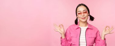 Farkındalık ve sağlık konsepti. Gülümseyen Koreli kız şık kıyafetler içinde meditasyon yapıyor, mantra dinliyor, zen 'de el ele tutuşuyor, barış pozu veriyor, yoga yapıyor, pembe arka planda duruyor.