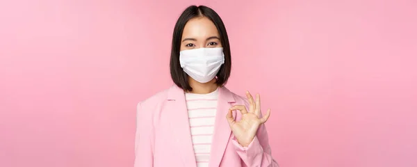Portrait einer asiatischen Geschäftsfrau mit medizinischer Gesichtsmaske, zeigt Okay-Zeichen, trägt Anzug, Arbeitsregeln während der Covid-19-Pandemie, steht über rosa Hintergrund — Stockfoto