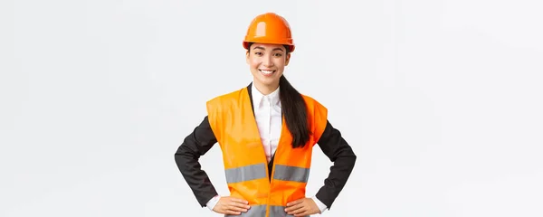 Exitosa joven mujer de negocios asiática, arquitecta en casco de seguridad, chaqueta reflectante, de pie confiada y decidida, sonriente asegurada, construyendo casas, posando sobre fondo blanco — Foto de Stock