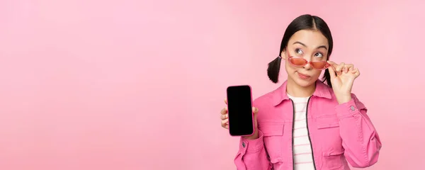 时尚的韩国女孩，戴着太阳镜、展示智能手机屏幕、手机应用程序接口或网站的年轻女性，站在粉色背景之上 — 图库照片