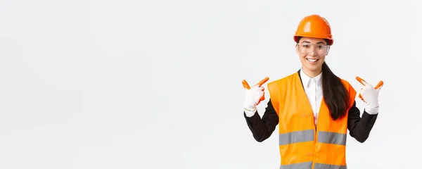 Sonriente confiada asiática jefa de ingeniería de construcción en casco de seguridad, guantes y gafas apuntándose a sí misma, notificar a seguir el protocolo seguro y usar ropa especial, fondo blanco — Foto de Stock