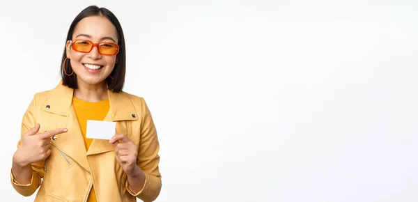 Retrato de hermosa chica asiática moderna en gafas de sol, sonriendo feliz, mostrando tarjeta de crédito, de pie sobre fondo blanco — Foto de Stock