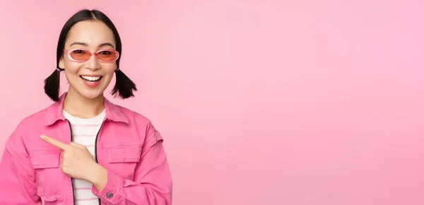 Retrato de sorrindo ásia menina no elegante roupa, óculos de sol, apontando dedo esquerdo, mostrando anúncio, banner, pé sobre rosa fundo — Fotografia de Stock