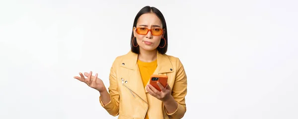 Porträtt av förvirrad asiatisk flicka i solglasögon, hålla smartphone, med hjälp av mobiltelefon och ser förbryllad, stående ledtråd över vit bakgrund — Stockfoto