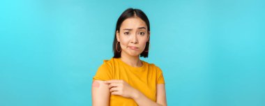 Covid-19 'dan aşı kampanyası. Üzgün ve hayal kırıklığına uğramış Asyalı kadın omzunu bandajlı ve asık suratlı, mavi arka planda dikiliyor.