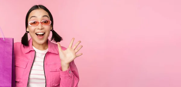 Vásárolni. Stílusos ázsiai lány napszemüveg, bemutató táska a bolt és a mosolygós, ajánló eladó promo a boltban, álló felett rózsaszín háttér — Stock Fotó