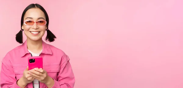 Портрет корейской девушки в стильных солнцезащитных очках с мобильным телефоном, с помощью приложения для смартфона, стоящей на розовом фоне — стоковое фото