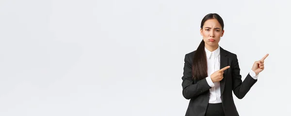 Disappointed or jealous female asian entrepreneur in suit complaining, pouting and pointing fingers right. Displeased saleswoman arguing, look offended or sad, standing white background — Stock Photo, Image