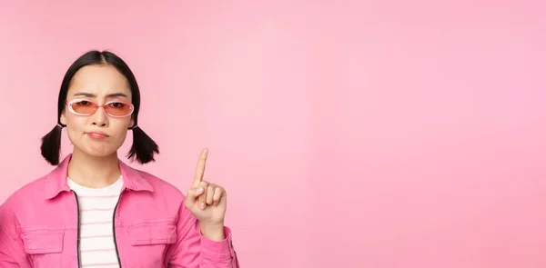 Primer plano de chica coreana con estilo, mirando perplejo, señalando los dedos hacia arriba, mostrando publicidad, de pie sobre el fondo rosa —  Fotos de Stock