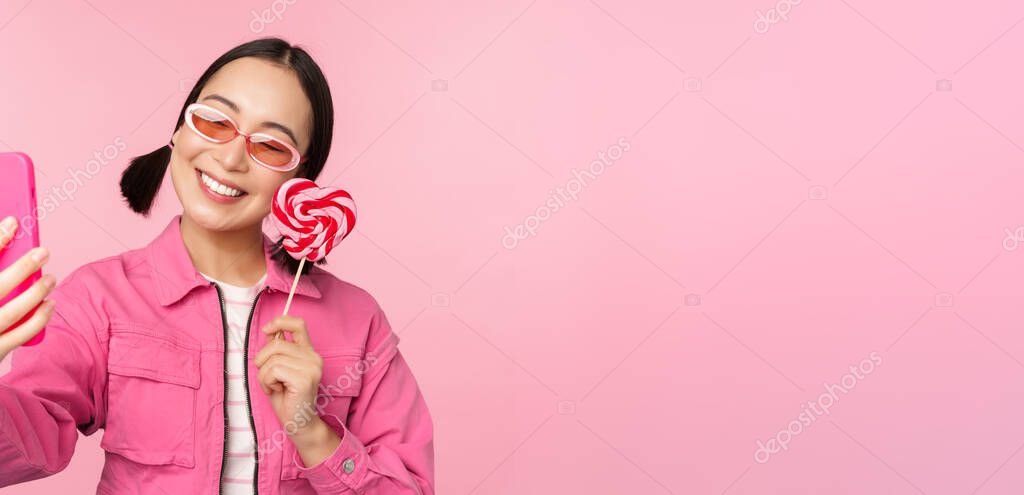 Portrait of stylish, happy asian girl taking selfie with candy, lolipop sweets and smiling, taking photo with mobile app, standing over pink background.
