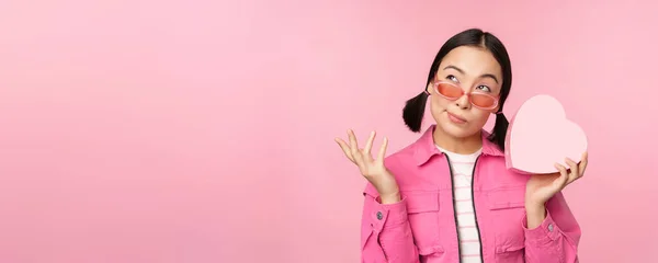 Εικόνα της κομψής Ασιάτισσας κοπέλας με γυαλιά ηλίου, μαντεύοντας τι είναι μέσα στο κουτί δώρου, δώρο σε σχήμα καρδιάς, στέκεται πάνω από ροζ φόντο — Φωτογραφία Αρχείου