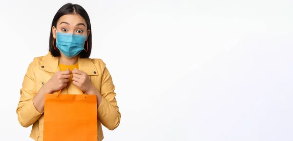Covid-19 e conceito de loja. Jovem mulher asiática elegante, vestindo máscara facial médica, segurando saco de compras, indo em shoppings durante a pandemia, fundo branco — Fotografia de Stock