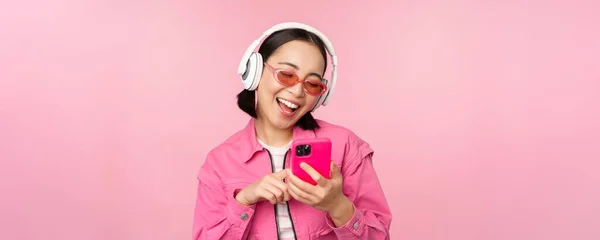 Elegante ragazza asiatica che balla con smartphone, ascolta musica in cuffia sull'app per cellulare, sorride e ride, posa su sfondo rosa — Foto Stock