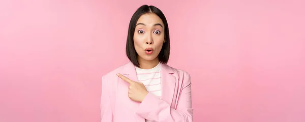 Retrato de mulher de negócios com rosto surpreso, apontando o dedo esquerdo, mostrando smth interessante, de pé sobre fundo rosa no terno de escritório — Fotografia de Stock