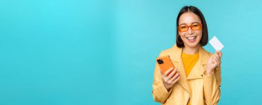İnternetten alışveriş. Güneş gözlüklü şık genç Asyalı kadın, kredi kartı gösteriyor ve akıllı telefon kullanıyor, internetten ödeme yapıyor, satın alıyor, mavi arka planda duruyor.