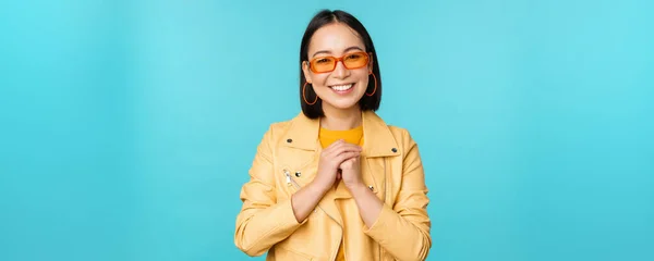 Porträt einer asiatischen Frau mit Sonnenbrille, die hoffnungsvoll, geschmeichelt, glücklich lächelnd vor blauem Hintergrund steht — Stockfoto