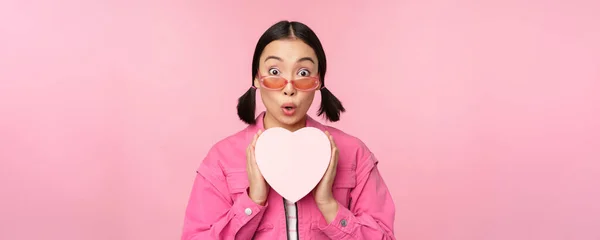 Linda menina asiática sorrindo feliz, mostrando caixa de presente do coração e olhando animado para a câmera, de pé sobre o fundo romântico rosa — Fotografia de Stock