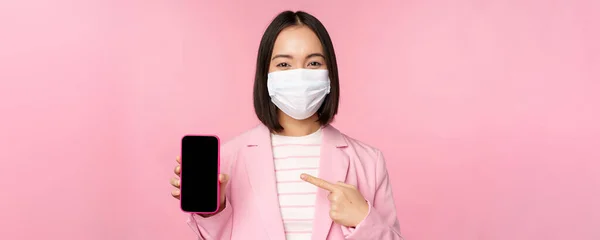 Smiling korean businesswoman in suit, wearing medical face mask from covid, pointing finger at mobile phone screen, standing over pink background — Stock Photo, Image