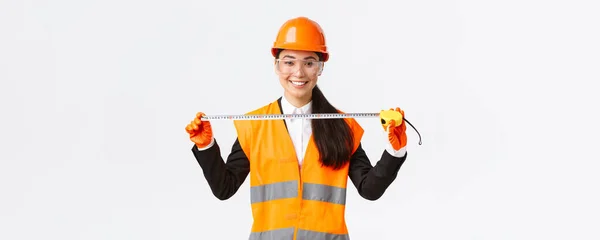 Pewna siebie profesjonalna kobieta azjatycki architekt układ pomiarowy, noszenie kasku bezpieczeństwa i jednolitego i trzymając miarkę taśmy, uśmiechnięty zadowolony, zadowolony z osiągniętego wyniku podczas budowy — Zdjęcie stockowe