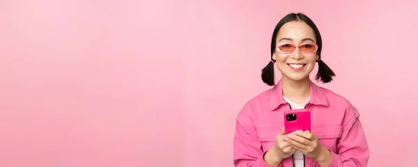 Porträtt av koreansk tjej i snygga solglasögon med mobiltelefon, med smartphone app, stående över rosa bakgrund — Stockfoto
