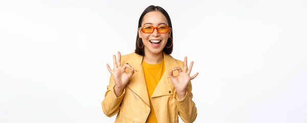 Entusiástico asiático menina em óculos de sol, mostra ok, ok assinar em aprovação, sorrindo e rindo, aprovar, recomendar smth, louvor e elogio, de pé sobre fundo branco — Fotografia de Stock
