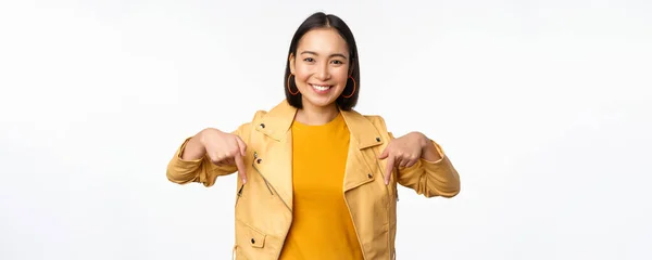 Mutlu gülümseyen Asyalı kızın portresi, parmakları aşağıda ve logoyu gösteriyor, pankartı gösteriyor, sarı ceketin içinde beyaz arka planda duruyor. — Stok fotoğraf