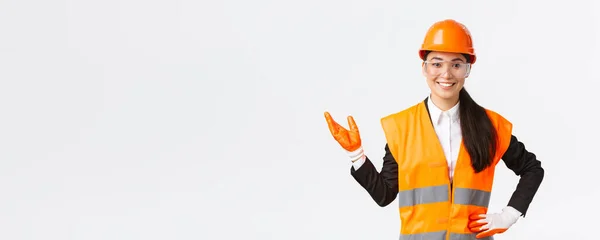 Glimlachend vrolijke Aziatische vrouwelijke bouwmanager introduceren haar verrassing voor investeerders, het dragen van een veiligheidsbril, helm en handschoenen wijzende hand links en op zoek naar trotse, witte achtergrond — Stockfoto